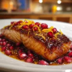 Glazed salmon fillet topped with pomegranate seeds and orange zest, served on a white plate with a glossy pomegranate sauce.